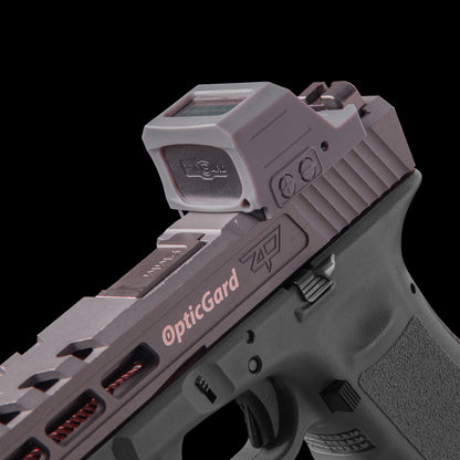 A close-up studio shot of a handgun with a mounted Gray OpticGard red dot Cover for Holosun 507CX3 against a solid black background. [ads-hide]