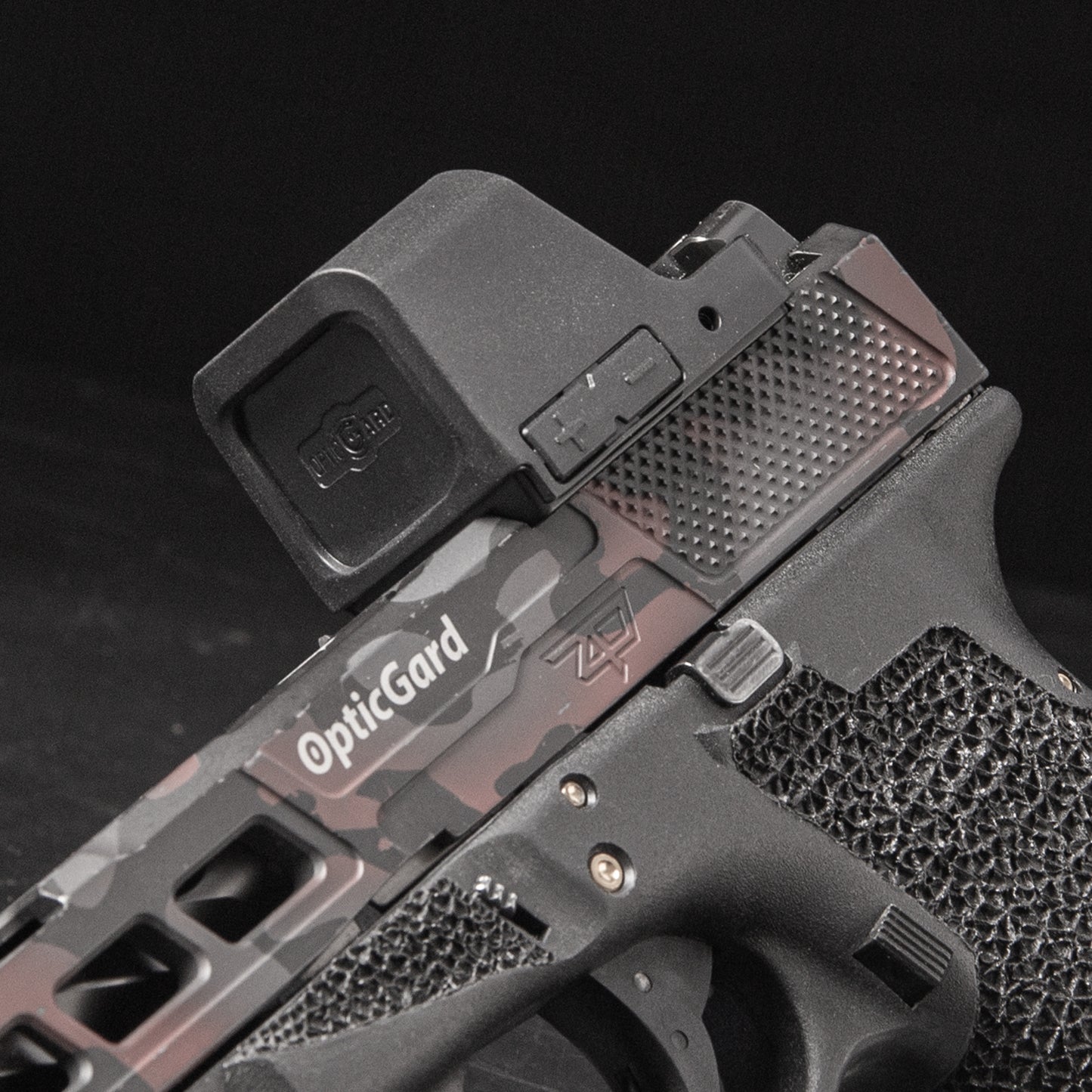 Close-up of a handgun with a mounted OpticGard red dot sight, featuring a dark gray and black camouflage pattern on the slide, against a black background. [ads-hide]