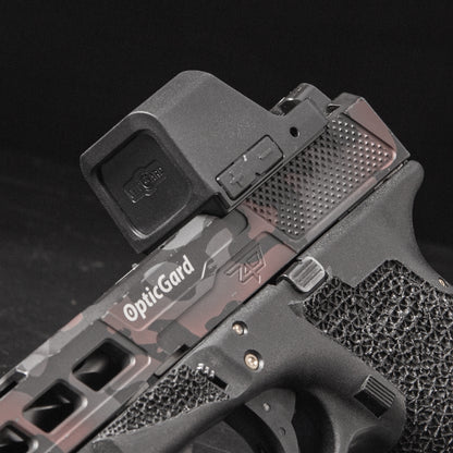 Close-up of a handgun with a mounted OpticGard red dot sight, featuring a dark gray and black camouflage pattern on the slide, against a black background. [ads-hide]