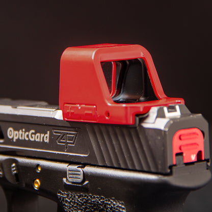 Close-up of a red OpticGard red dot cover on a black handgun against a dark background. [ads-hide]