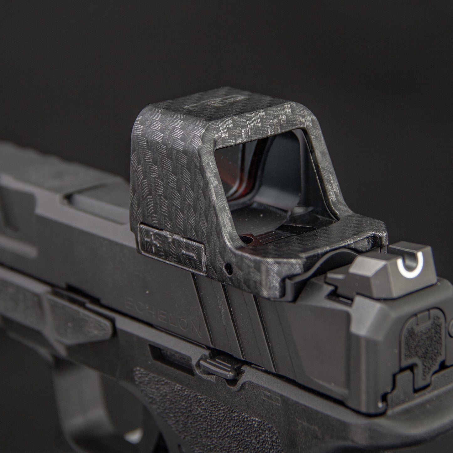 Close-up of a carbon-fiber-patterned  red dot cover mounted on a black handgun against a dark gray background. [ads-hide]