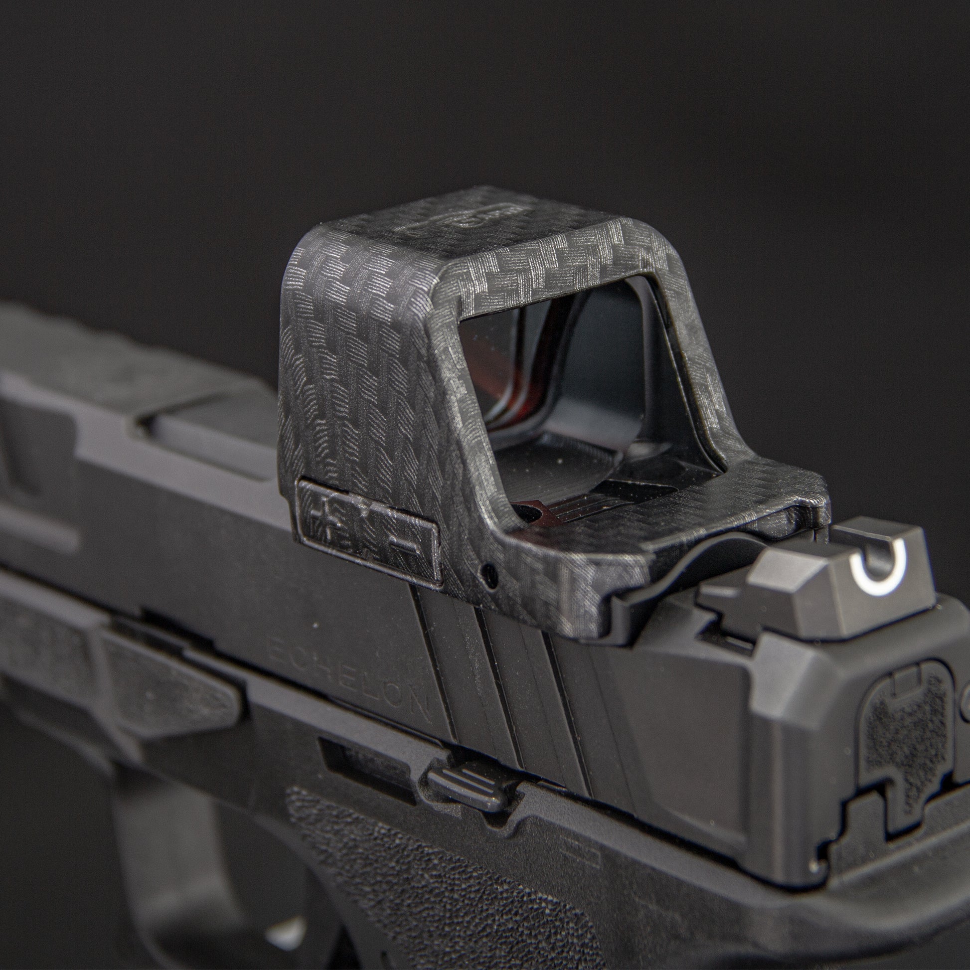 Close-up of a carbon-fiber-patterned  red dot cover mounted on a black handgun against a dark gray background. [ads-hide]