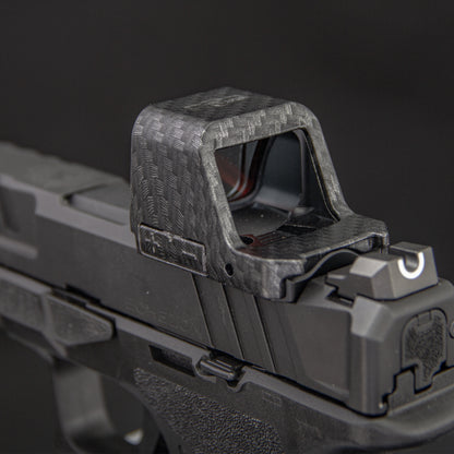 Close-up of a carbon-fiber-patterned  red dot cover mounted on a black handgun against a dark gray background. [ads-hide]