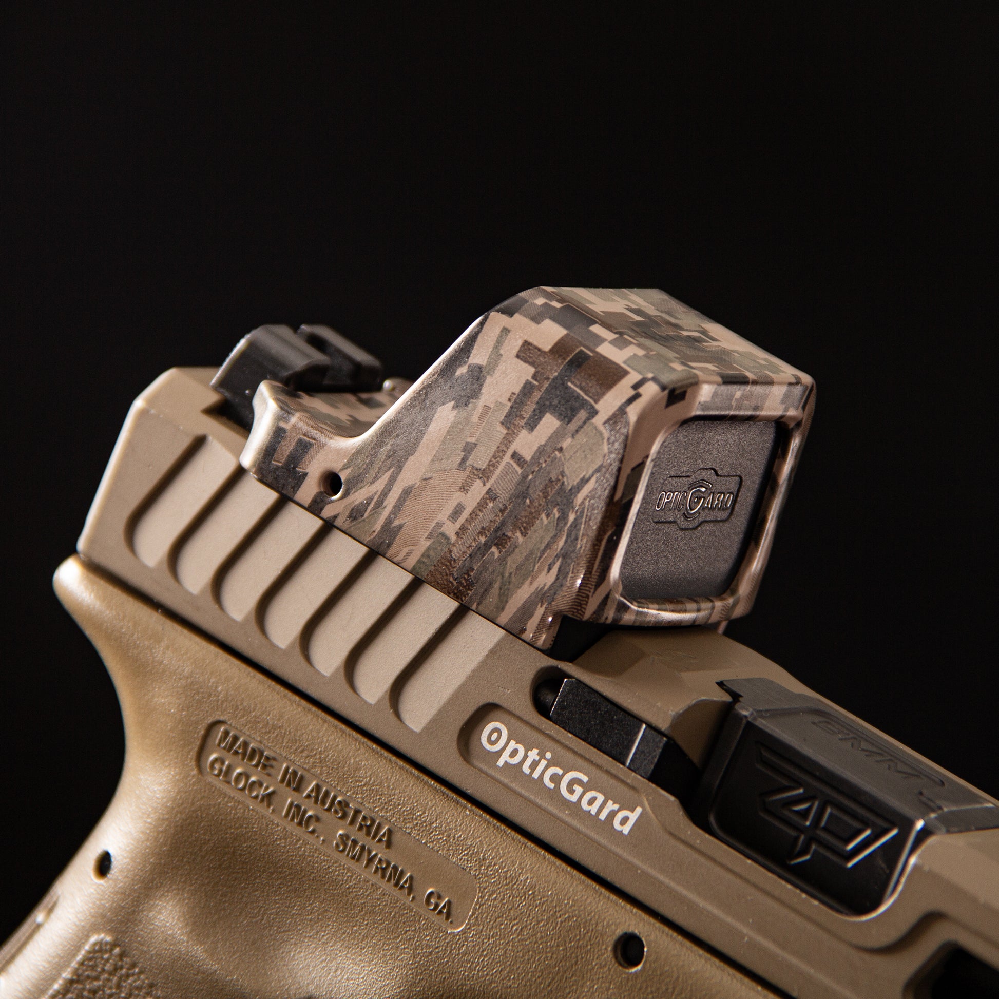A close-up of a FDE camo OpticGard red dot sight cover with a digital camouflage pattern mounted on a tan handgun against a solid black background. [ads-hide]