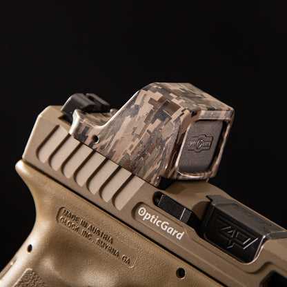 A close-up of a FDE camo OpticGard red dot sight cover with a digital camouflage pattern mounted on a tan handgun against a solid black background. [ads-hide]